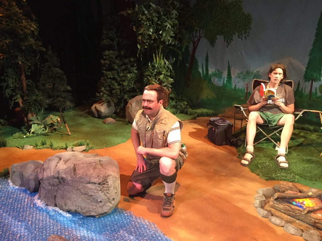 Two male campers relax in their campsite. One sits in a camp chair and reads a book, while the other knells down to contemplate a fake river made of blue bubble pop.