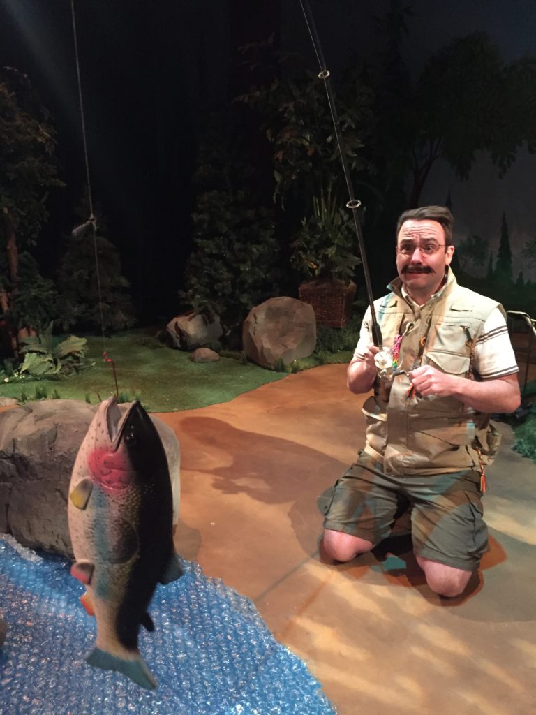 A male camper wearing a mustache and glasses looks worried as he contends with the fake fish that he’s caught.