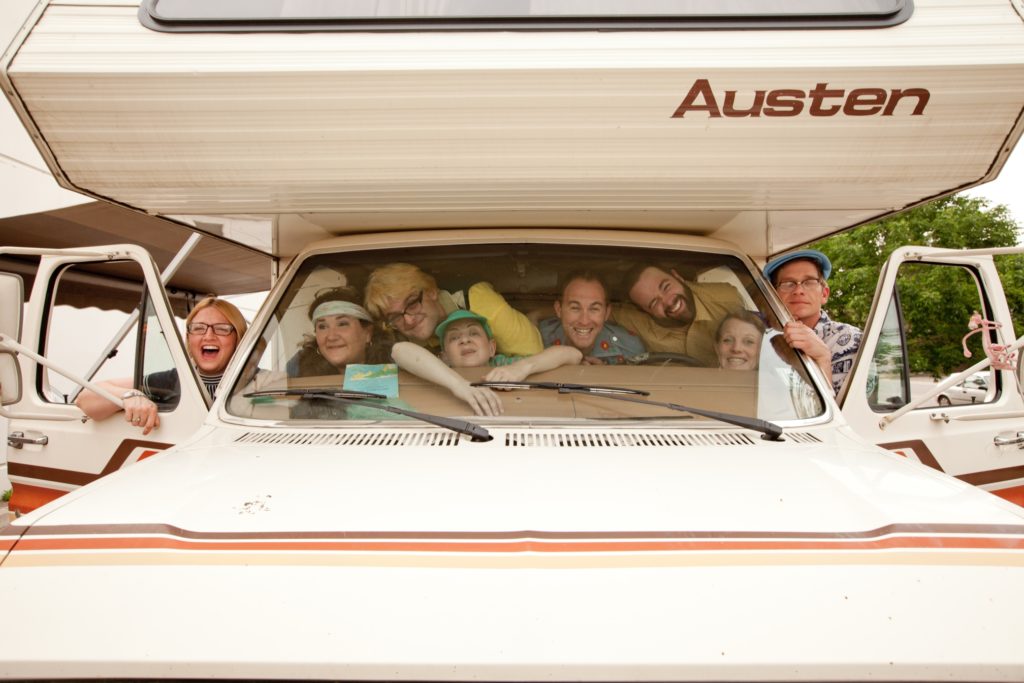 Six people are jammed into the front window of an old camper van, with two more people standing on the sides of the van, in the open doors. Most people are smiling, despite the tight squeeze.