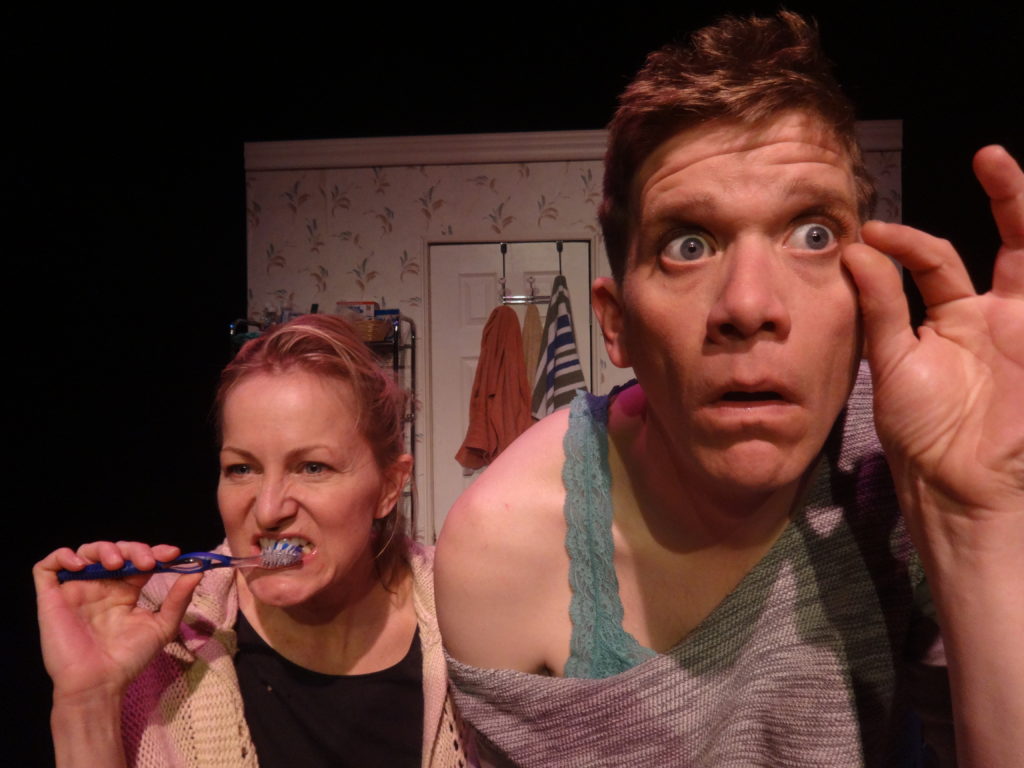 Two people stand at the bathroom sink. While looking in the mirror one brushes their teeth while another checks their eyelashes.