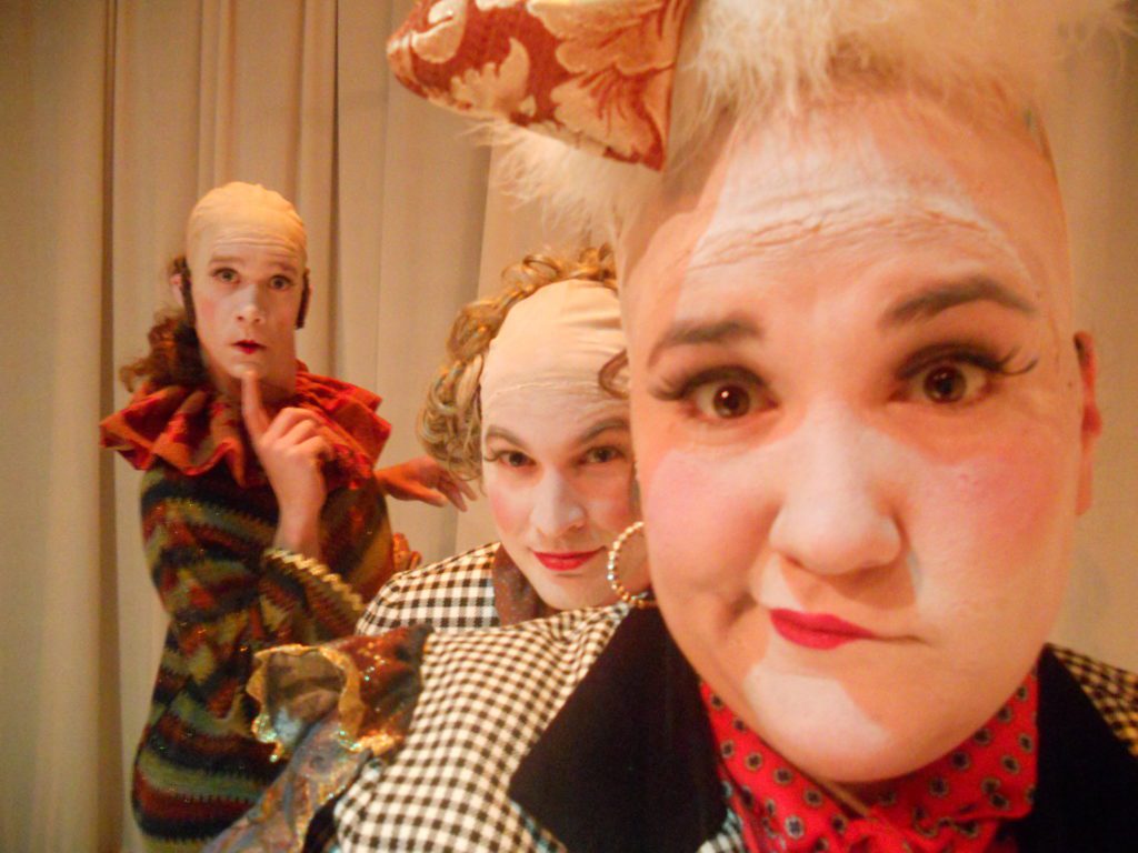 In the foreground a clown that has a puff of white feathers for hair looks into the camera like they don’t know what’s going on. The clown right over their shoulder looks as if they are sneaking into the picture. The clown in the background holds a finger to their chin as if they are posing for senior portrait.