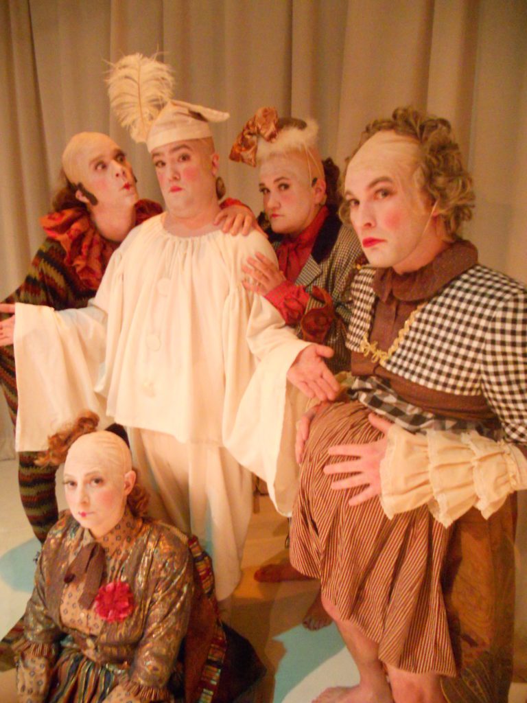 A group of four clowns in multi-colored and patterned clown outfits surround another shrugging clown, who wearing all white.