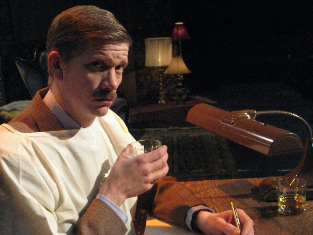 A man with a large mustache, dressed in a suit, and wearing a hospital gown sits at his desk. The man holds a glass of whiskey while deciding what to write. His expression is wry…or maybe just serious.