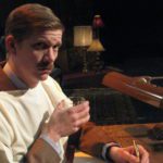 A man with a large mustache, dressed in a suit, and wearing a hospital gown sits at his desk. The man holds a glass of whiskey while deciding what to write. His expression is wry…or maybe just serious.