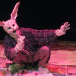 A large rabbit wearing a ratty robe and bunny slippers squats on a floor covered with newspapers. The rabbit holds a microphone and sings.