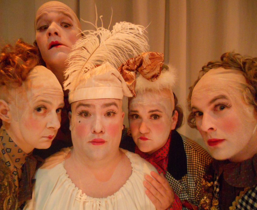 Five people dressed as clowns with bald caps and multi-patterned clothes look into the camera inquisitively.