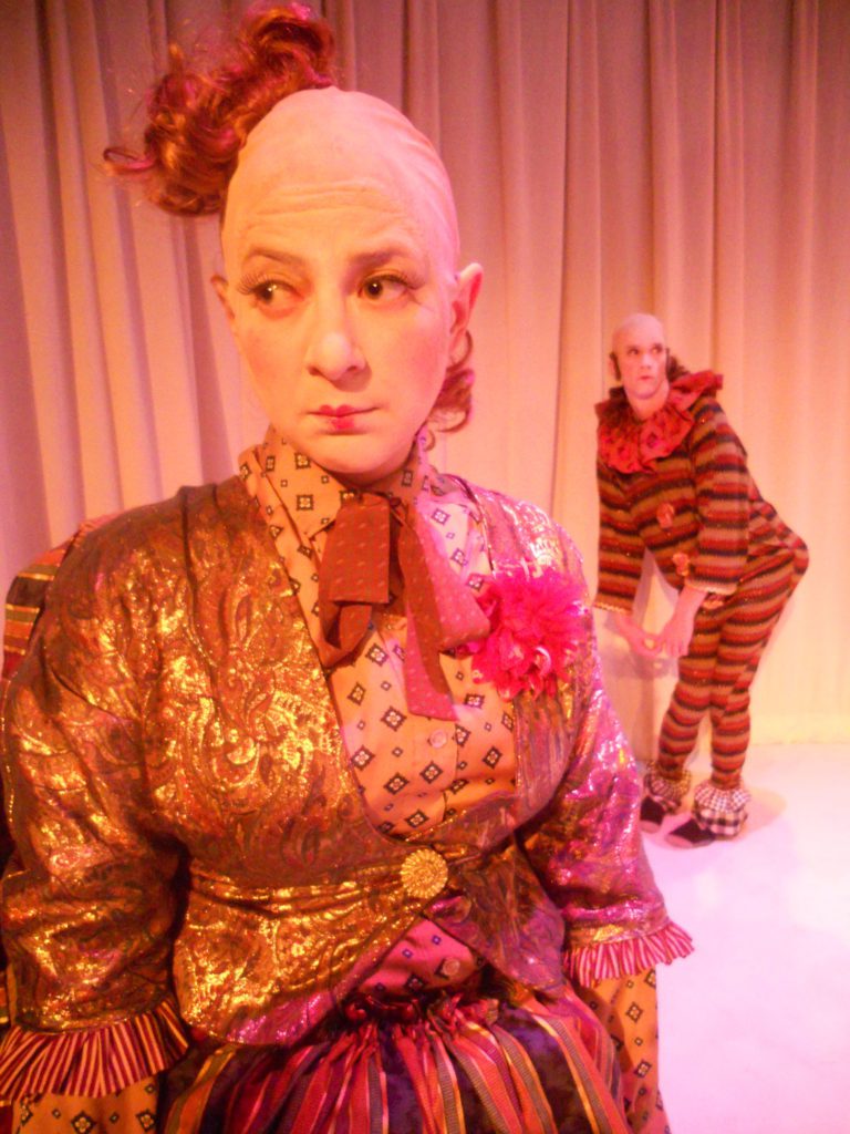 A clown with a poof of fabric for hair, looks off cloyingly in the foreground, while another clown behind them sticks out their big fake butt looking as if they had just farted.