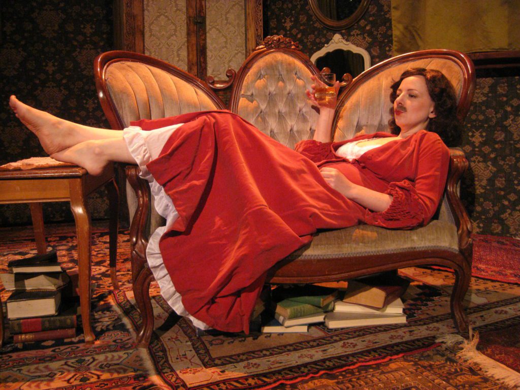 A mustached woman, wearing a red velvet gown, reclines on a turn of the century love seat. Her bare feet dangle above a piano bench set to the right of the love seat. The woman holds a glass of whiskey and puckers her lips. Books are stacked in separate piles on the floor.