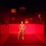 A woman strikes a strong pose, arms akimbo, center stage. The room she stands in is entirely red and decorated with her portraits.