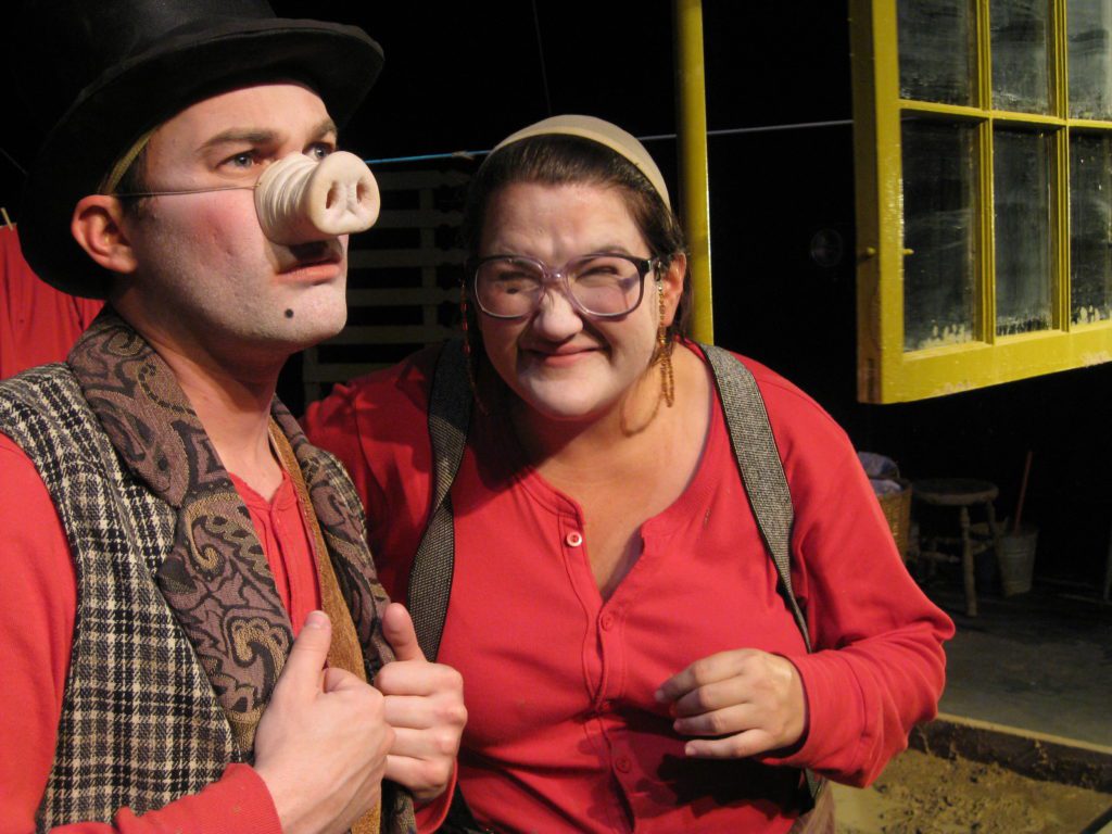 A man in a pig nose, red long johns, and a vest looks out, while a woman in red long johns, suspenders, and glasses smiles at him. Behind them a pea green window hangs.
