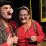 A man in a pig nose, red long johns, and a vest looks out, while a woman in red long johns, suspenders, and glasses smiles at him. Behind them a pea green window hangs.
