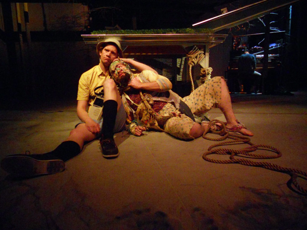 A man dressed in rags and tied with thick rope to a platform is comforted by a man carrying a tape recorder who is dressed for summer holiday.