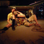 A man dressed in rags and tied with thick rope to a platform is comforted by a man carrying a tape recorder who is dressed for summer holiday.