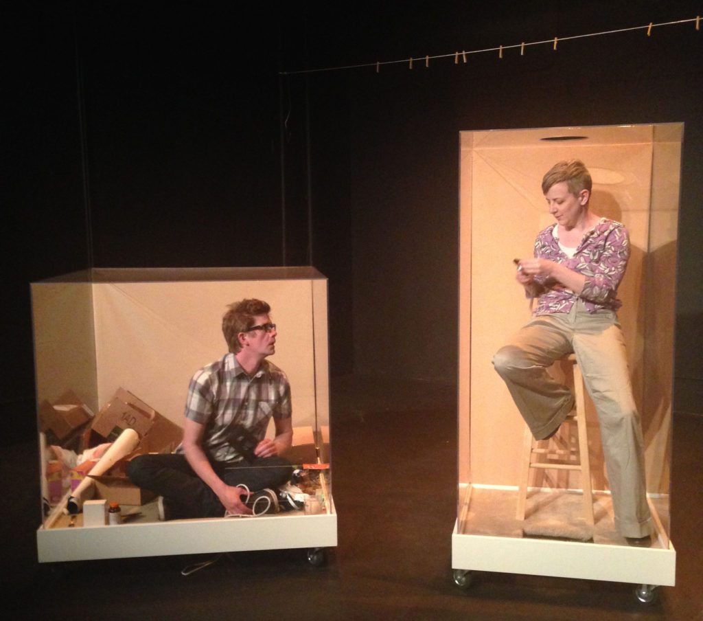 Two people talk to each other from inside plexiglass boxes. On the left is a man sitting crosslegged in a short, cluttered box. On the right is a woman sitting on a stool in a narrow box. Above is an empty clothing line.