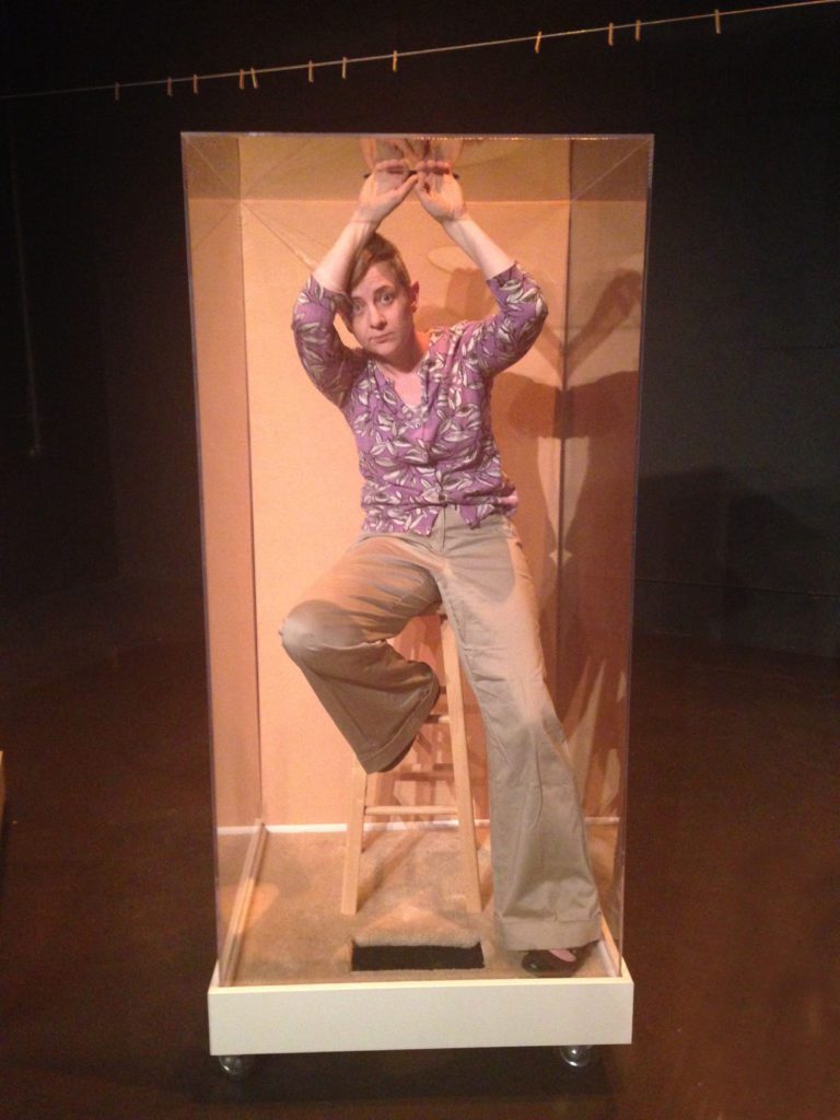 A woman with short hair sits on a stool in a plexiglass box. She has one leg up on the school and has both hands above her head, holding onto the rim of a hole in the top of the box. Above the box is a clothing lines with only clothes pins on it.