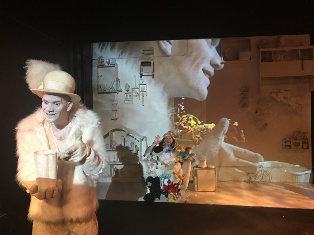 A man dressed all in white with a fur coat and hat with a large feather in it smiles creepily. He is holding a large white container. Behind him, on the wall, is a projection of his own profile. In the distance, with the projection on her, is a woman covered in stuffed animals. She is waving.