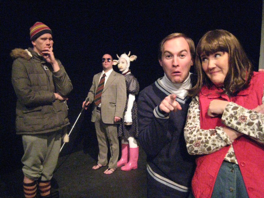 A couple smile goofily for the camera. Behind them are a disheveled man dressed for winter, a man in a suit and sandals holding a white cane, and a cow in a polka dot dress and rain boots.