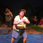 A woman in full camp counselor regalia stands posed in front of a fake river made of blue bubble pop. Behind her two male campers stand holding one another. A Resusci Annie sits alone in a camp chair to the left.