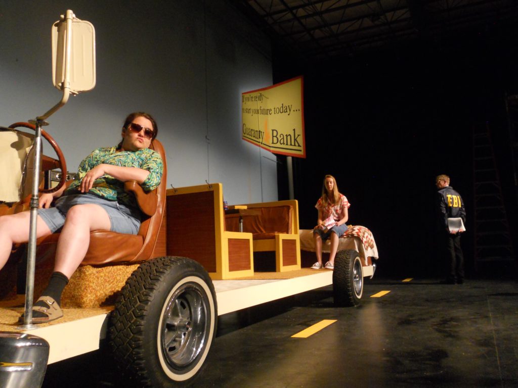 Just the insides of an RV sit on a platform with wheels. Sitting in the RV are two people, one lounging in the driver's seat, the other sitting in the back on the bed. In the background in a road sign for a bank and an FBI officer with his back turned.