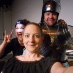 Two women, and a man dressed in a suit of armor poise for a selfie. One of the woman is wearing a head lamp and making the devil horns rock-and-roll sign with her hands.