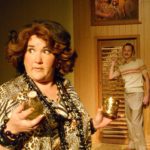 A woman with a large bouffant hairdo and a leopard-print caftan holds a bottle and glass. Behind her, a man with a mustached stands in front of a slatted wooden door with a family portrait hanging above.