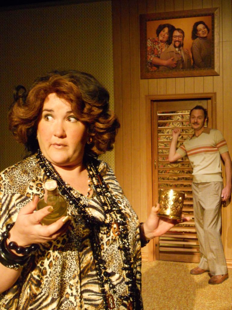A woman with a large bouffant hairdo and a leopard-print caftan holds a bottle and glass. Behind her, a man with a mustached stands in front of a slatted wooden door with a family portrait hanging above.