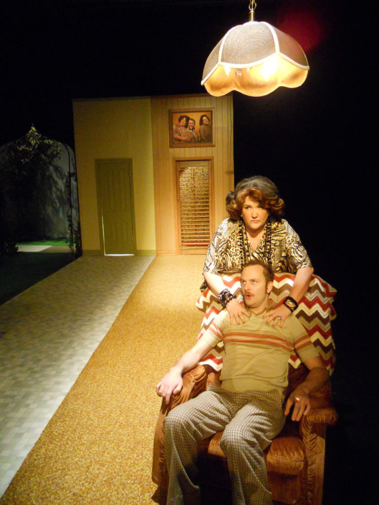A man sits in a Barca lounger with a 1970s crocheted throw over the back. A woman with big hair stands behind him, rubbing his shoulders. Above them is a lamp. In the background is a wall with three sections: the living room, kitchen, and outside.