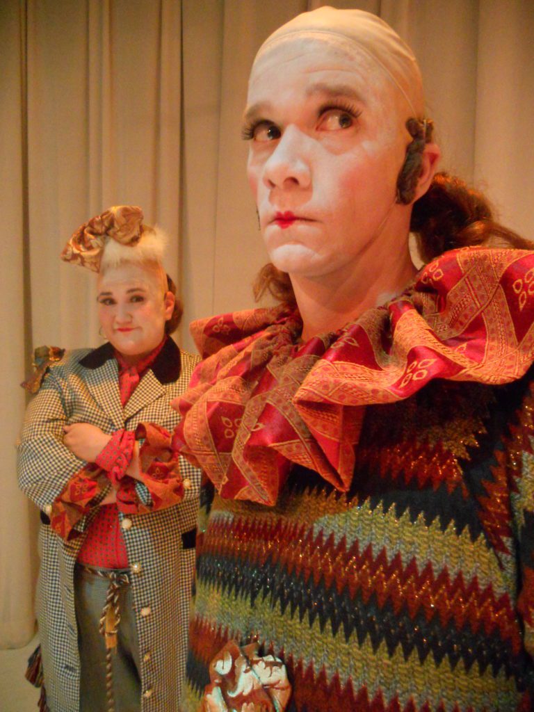 In the foreground, a clown wearing a huge ruffle collar looks off in the distance while another clown in the background looks into the camera with a smile on their face and arms crossed.