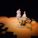 A life-size puppet of Tommy Lee Jones sits at a diner table that is surrounded by a yellow and white checkered floor surrounded by darkness. A waitress stands next to the table as if she is about to take his order.