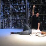 A minister kneels, reaching skyward, and places his hand on the chest of a man laying on the floor. Behind the two men is a giant wall made of jars filled with sundry objects.