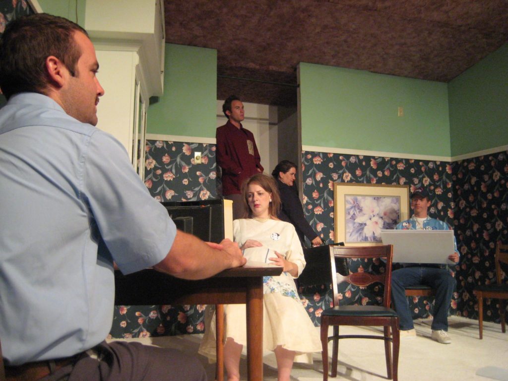 The setting is an upside down room, with pink carpet on the ceiling and flowered wallpaper. Five people crowd into the room, each doing their own activity.