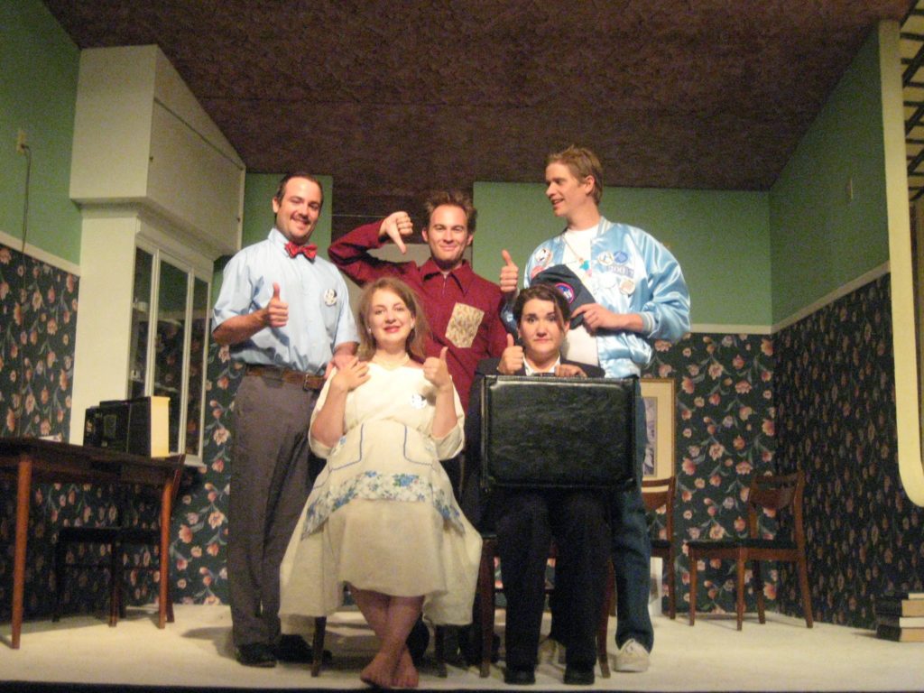 Four smiling people give the camera a thumbs up while one smiling man gives a thumbs down. They are grouped together in the middle of an upside down room.