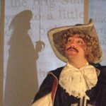 A man with a mustache and curly blond hair stands regally in a musketeer outfit. In the background there is a silhouette of another musketeer holding a sword, surrounded by projected text.