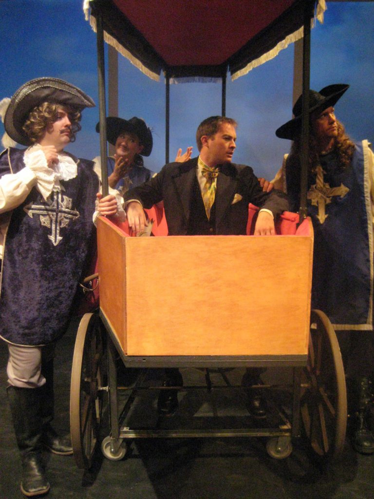 A man sits in a carriage that is shaped like a coffin. Surrounding him are three people dressed as musketeers.