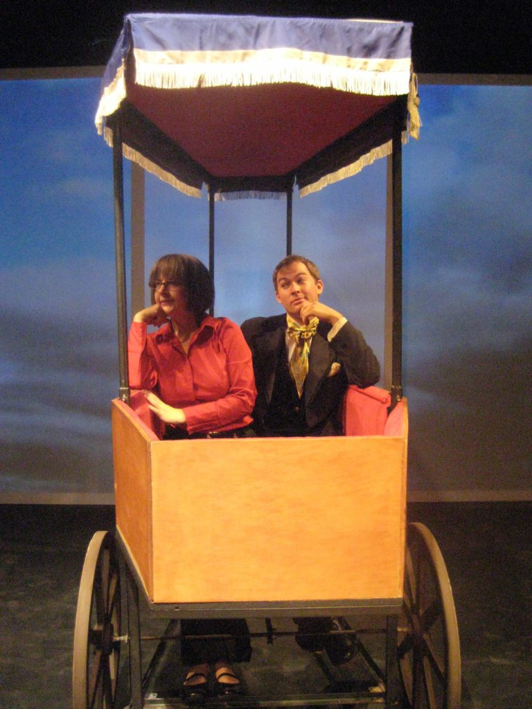 A modern woman and a man from the 1800s sit in a carriage shaped like a coffin. Behind them, an image of the sky is projected on a giant screen.
