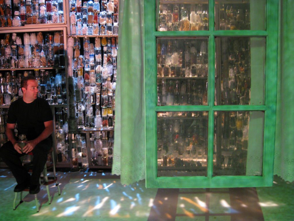 A man dressed in black sits on a stool holding a jar. Behind him light shines through a wall made of jars filled with sundry objects. In the foreground a window with curtains, bathed in green light, hangs in the air.