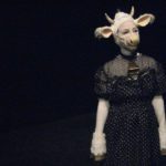 A woman dressed as a white cow in a polka dot dress stands, surrounded by darkness.