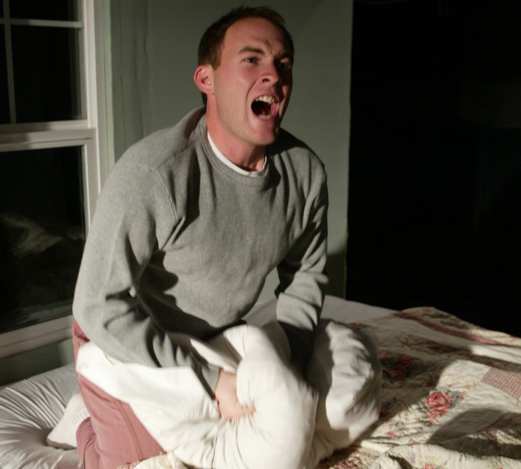 A blonde man in a grey sweatshirt, white socks and purple pajama bottoms is sitting up on his knees on a bed and yelling angrily outward and to the right. He is clenching in front of his lap a white flower-patterned blanket. Behind him is a white wall with a partially open double-hung window.