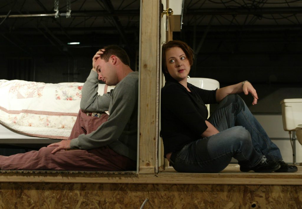 A cross section view of a door with a man and woman sitting on either side with their backs against the door. The woman is on the right side of the door facing to the right with her head turned forward as if she is speaking to the man behind her on the other side of the door. The man is looking at the floor, upset.