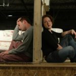 A cross section view of a door with a man and woman sitting on either side with their backs against the door. The woman is on the right side of the door facing to the right with her head turned forward as if she is speaking to the man behind her on the other side of the door. The man is looking at the floor, upset.