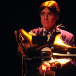 Close-up of an unhappy looking man in stark lighting. He has many pairs of ice skates draped over his shoulders.
