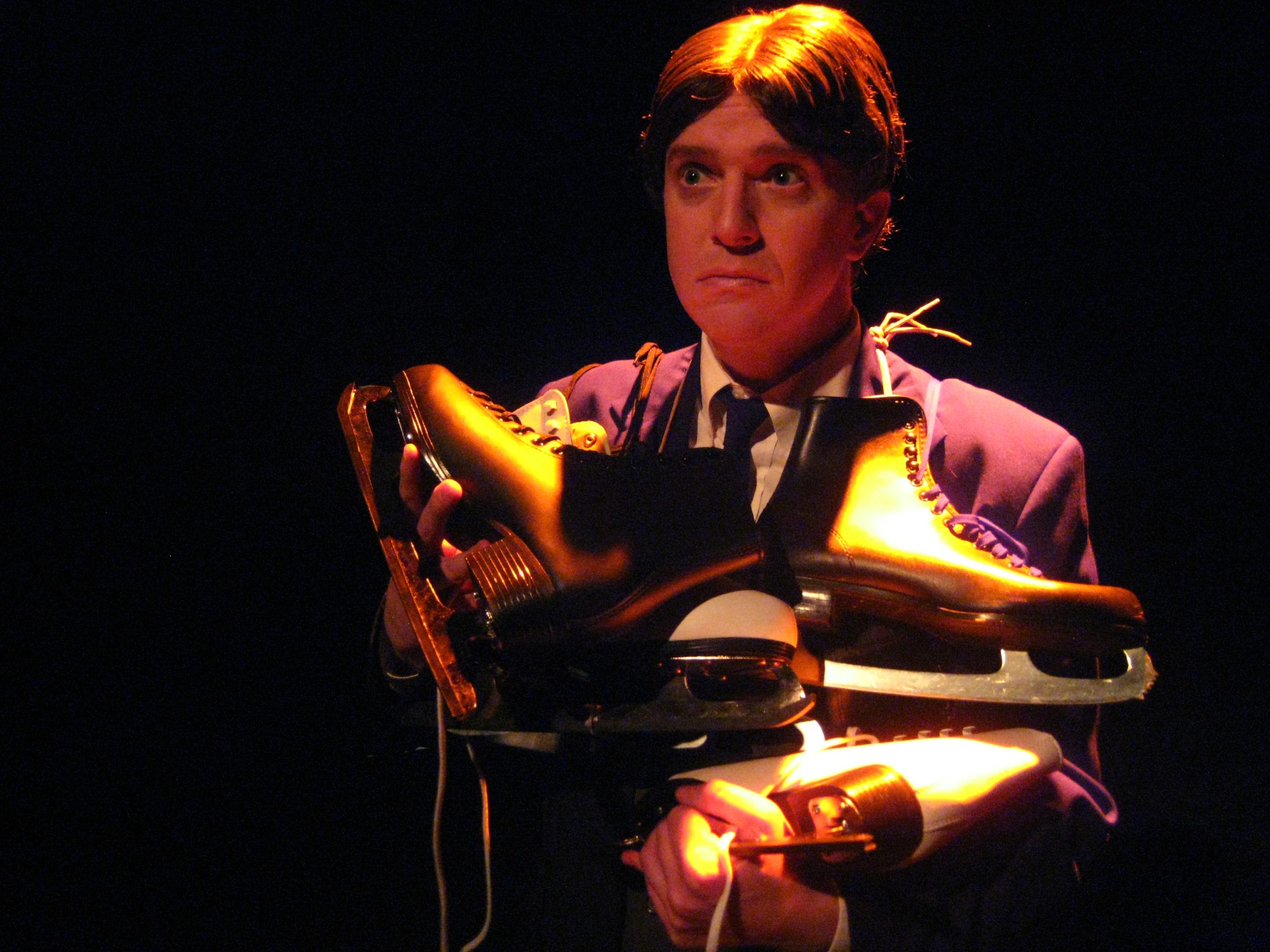 Close-up of an unhappy looking man in stark lighting. He has many pairs of ice skates draped over his shoulders.