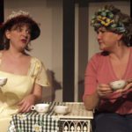 Two women holding teacups gossip at a small cafe table. One wears a hat, the other is in curlers.