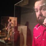 Close-up on an unhappy bearded man holding a telephone. He is behind plexiglass. In the distance are three other people sitting in large plexiglass boxes.