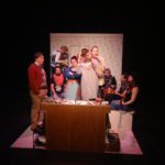 A group of people are crowded into a bathroom. A band with instruments sits in the bath tub. Two people, seemingly entwined, attempt to brush their teeth in the foreground.