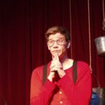 A man in a red top, black suspenders, and glasses looks out inquisitively. Behind him are several hanging buckets in front of a red curtain.