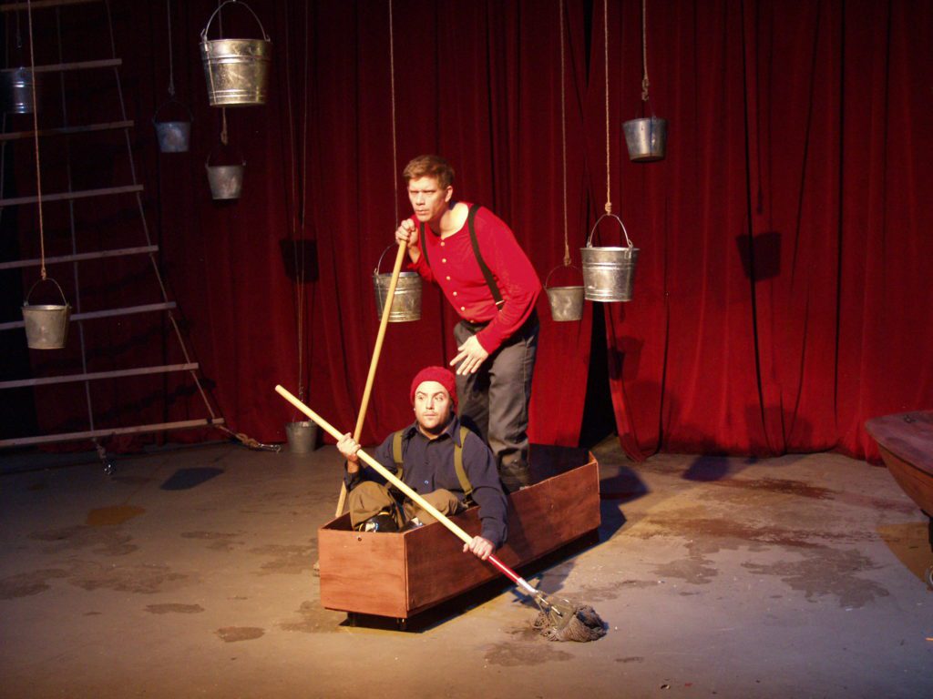 Two men, one seated and one standing, are “rowing” in a wooden coffin, using mops as oars. In the background is a red curtain, a rope ladder, and several hanging buckets.