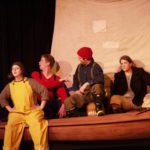 Three sailors sit in a small sailboat. A fourth sits on the edge of the boat, wearing bright yellow waders