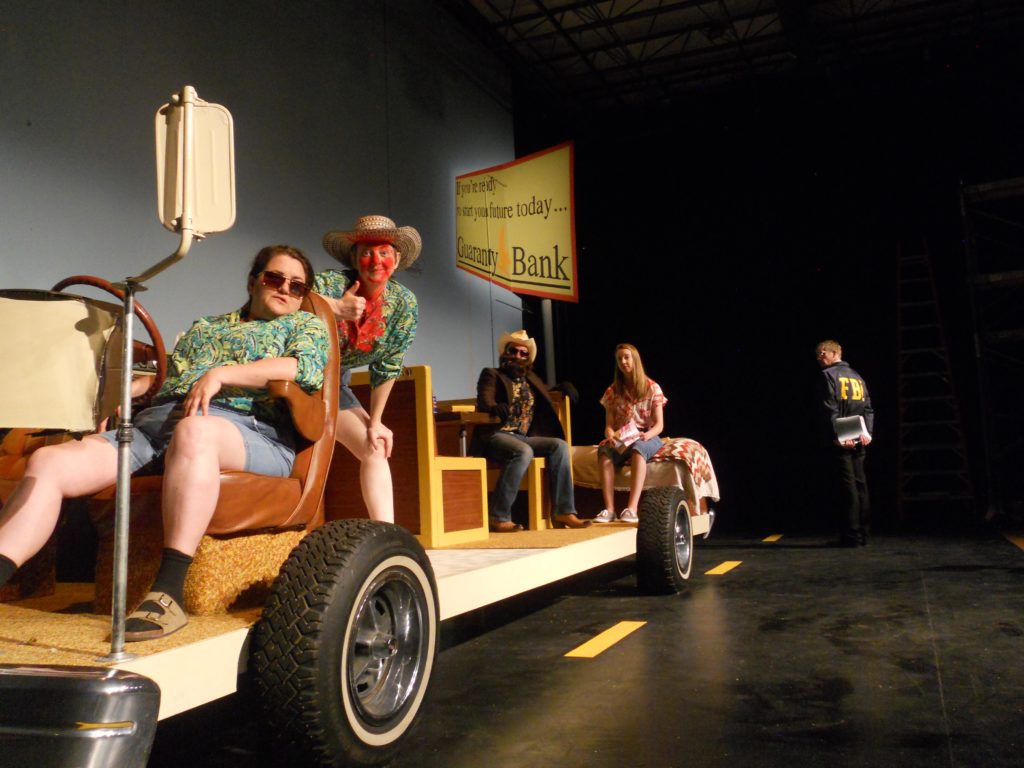 Just the insides of an RV sit on a platform with wheels. Sitting in the RV are two people, one lounging in the driver's seat, the other sitting in the back on the bed. In the background in a road sign for a bank and an FBI officer with his back turned.