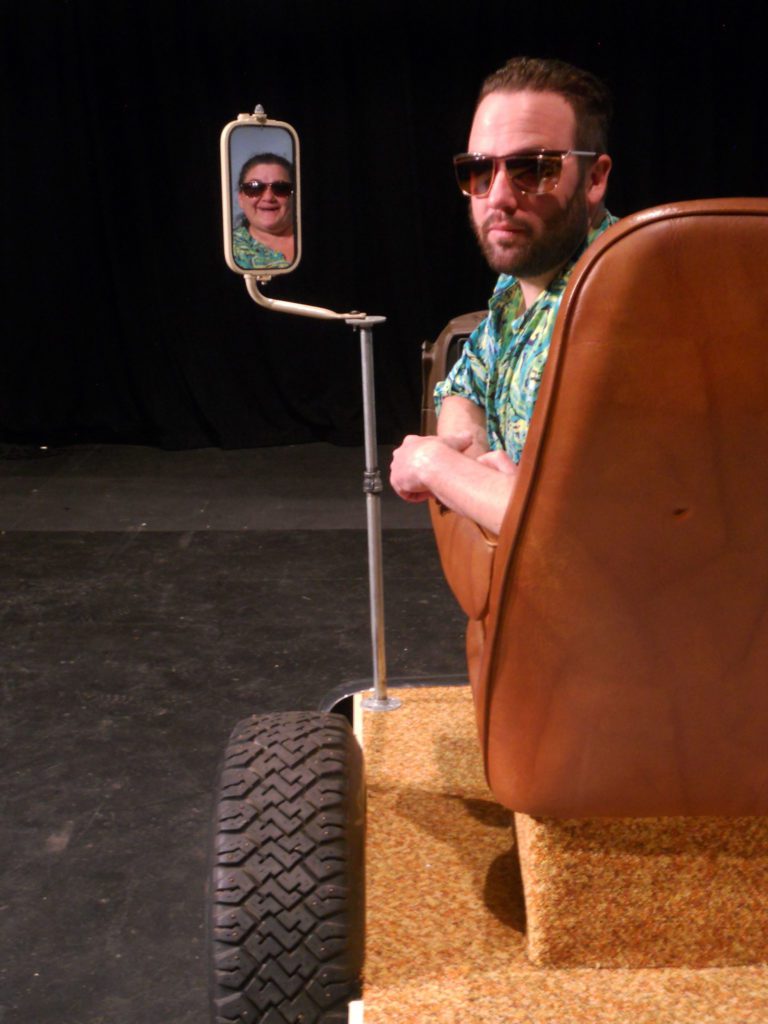 A man with sunglasses and a brightly colored shirt sits in a brown driver's seat, twisting to stare seriously at the camera. Next to him, in a driver's side mirror, a woman in the same sunglasses and shirt is smiling.